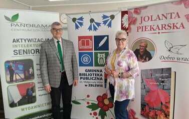 Pisarka i Jej mąż stoją obok siebie na tle baner&oacute;w promujących Centrum Szkolenia Panorama, Gminną Bibliotekę Publiczną w Zblewie oraz książki Jolanty Piekarskiej.