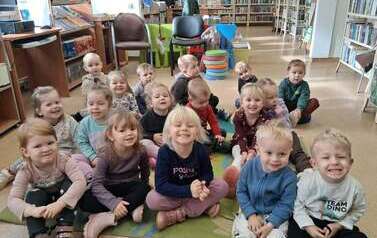 Dzieci siedzą na dywanie i słuchają bajki kt&oacute;rą czyta bibliotekarka