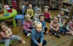 Dzieci siedzą na siedziskach i słuchają bajki, którą czyta im bibliotekarka.
