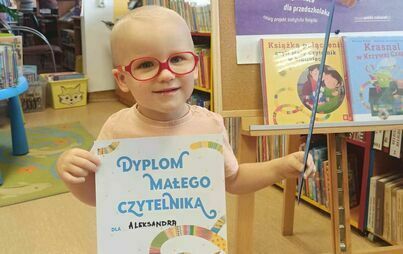 Chlopiec w blond wloskach i czerwonych okularach trzyma swój dyplom Małego Czytelnika