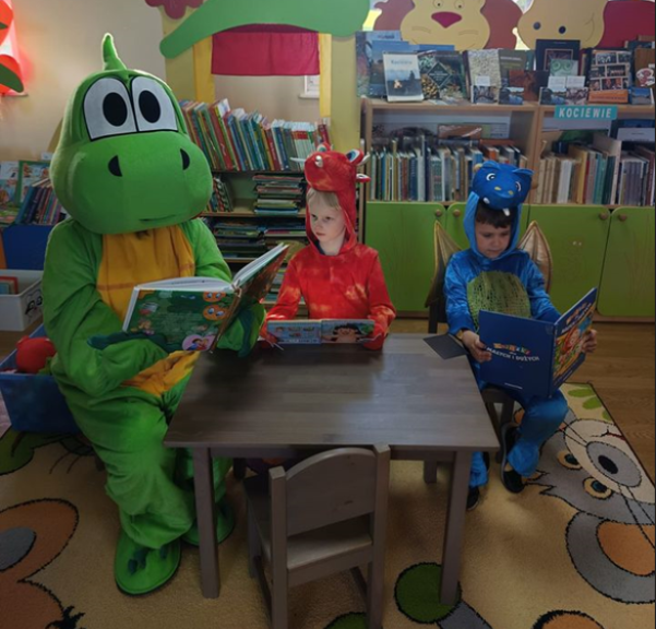 Smok duży i&nbsp;dwa małe siedzą przy stoliku i&nbsp;czytają bajki.