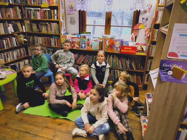 Dzieci siedza na dywanie i słuchają czytanej bajki przez bibliotekarkę.