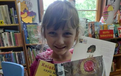 Dziewczynka w blond włosach uśmiechając się pokazuje swoją wyprawkę czytelniczą.