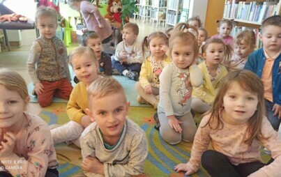 Dzieci siedzą na dywanie i słuchają bajki, którą czyta im bibliotekarka.