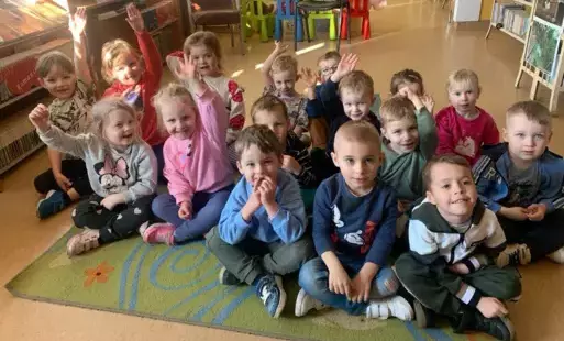 Dzieci siedzą na dywanie, niektóre z nich podnosi rękę w celu zabrania głosu