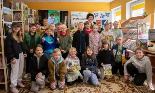 Dzieci stoją w grupie z Anną Sakowicz. Przednie dzieci siedzą na dywanie i niektóre z nich trzymają zakpione książeczki z serii - Poprańcy