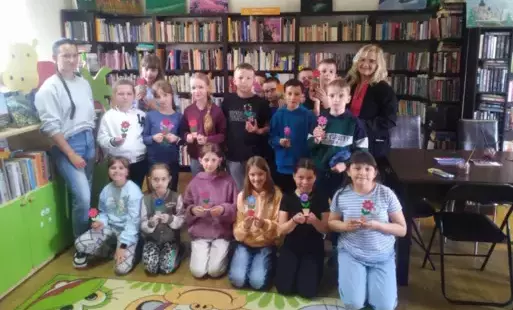 Dzieci stoja w grupie ze swoją Panią i Panią, która im czytała bajkę. Trzymają w dłoniach drewniane małe kwiatki, które pomalowali na Dzień Matki