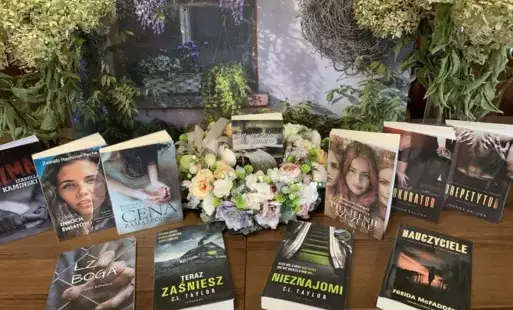 Nowe książki leżą na stole z widocznymi tytułami, po środku leży wianek z kwiatów z książką w środku. W tle jest dom obrośnięty fioletowym bzem.