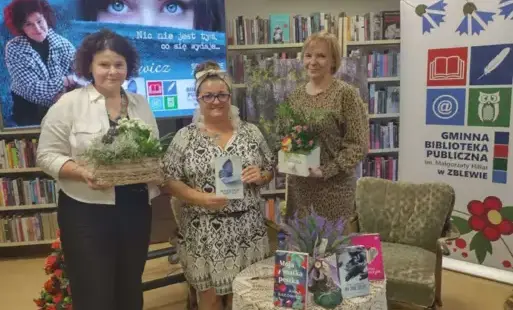 Stoją trzy Panie: Pani Sakowicz, Pani kierownik biblioteki i Pani prowadząca spotkanie. Trzymają przystrojone kwiaty w pudełkach i kierowniczka trzyma książkę pisarki.