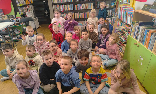 Grupa dzieci siedzi na dywanie i patrzy w stronę bibliotekarki prowadzącej zajęcia