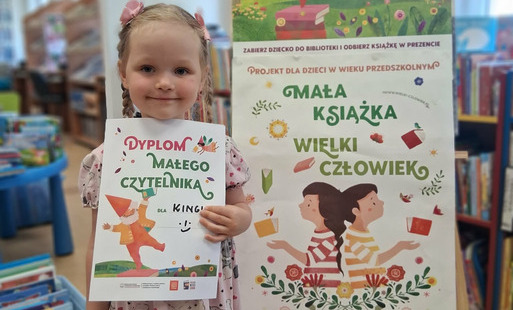 Dziewczynka w blond warkoczykach pokazuje swój dyplom, obok stoi tablica z plakatem dotyczącym projektu Mała Książka Wielki Człowiek