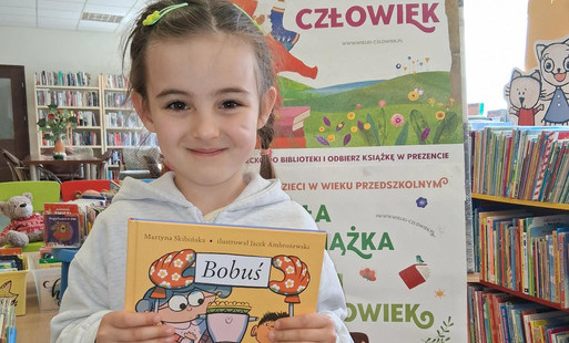 dziewczynka pokazuje swoja nową książkę pt Rita i koń. Za nią stoi tablica z plakatami projektu Mała Książka Wielki Człowiek