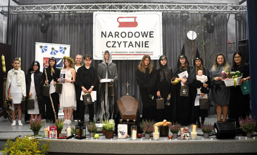 Młodzież stoi na scenie z kierowniczką biblioteki w strojach nawiązujących do dramatu. w tle napis "Narodowe Czytanie", a scenę ozdabiają dekoracje i rekwizyty nawiązujące do obrzędu dziadów.