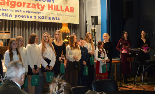 Grupa dziewcząt i chłopców oraz opiekunka stoją do wspólnego zdjęcia podczas uroczystości. Trzymają torby z upominkami. W tle baner z napisem Rok Jubileuszowy Małgorzaty Hillar