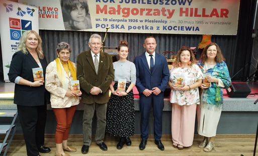 Przed sceną stoi 5 kobiet i dwóch mężczyzn pozując do zdjęcia. Kobiety trzymają książkę o Małgorzacie Hillar