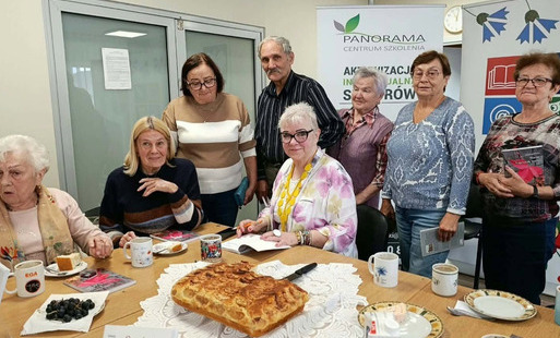 Uczestnicy spotkania siedzą i stoją wokół stołu z poczęstunkiem. Na stole znajduje się ciasto, owoce, fliżanki i talerzyki. W tle widoczny baner Centrum Szkolenia Panorama.